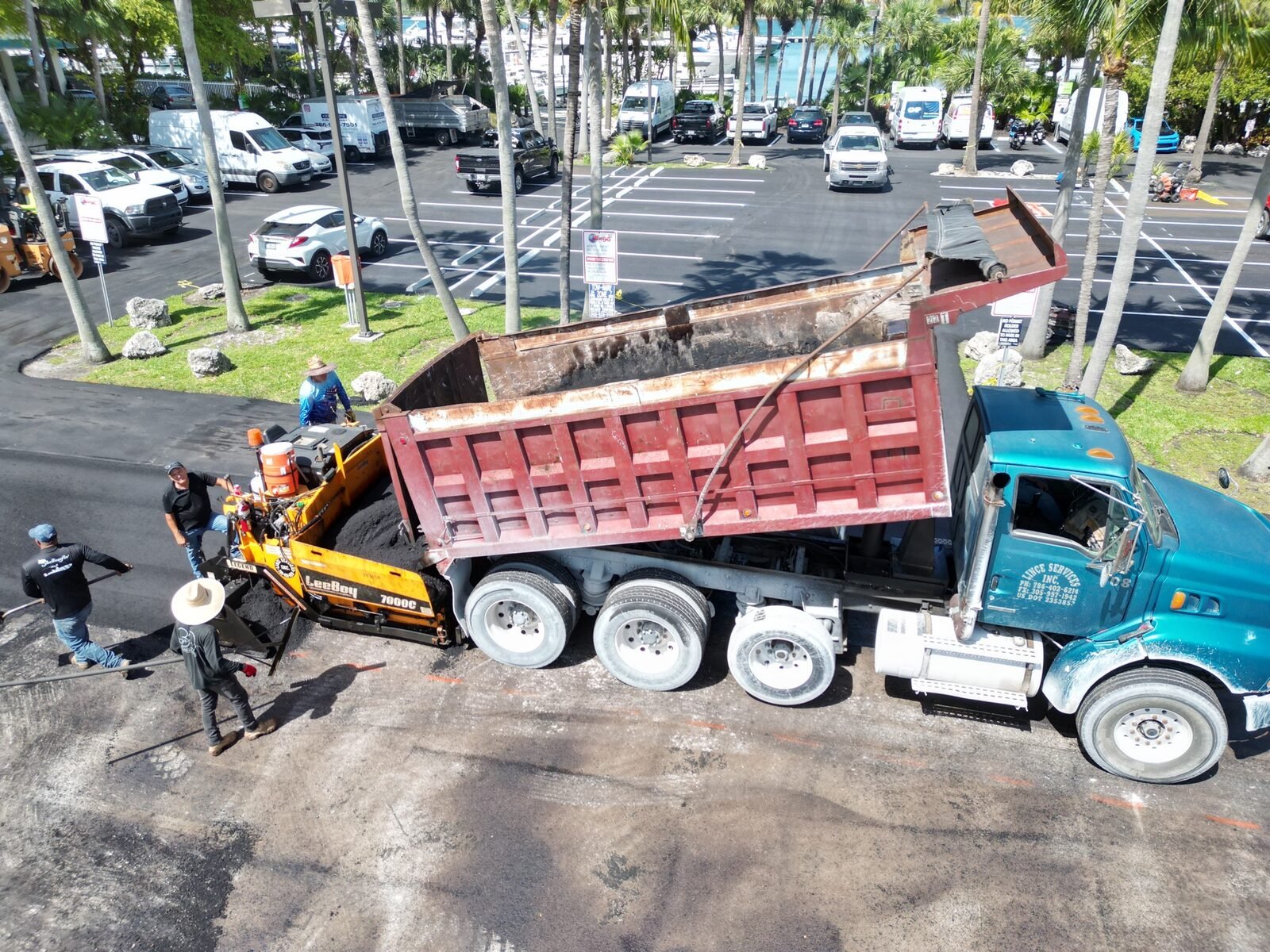 Souffront crew resurfacing a South Florida commercial parking lot — a dump truck charges the asphalt paver hopper while crew members finish the edges with hand rakes.